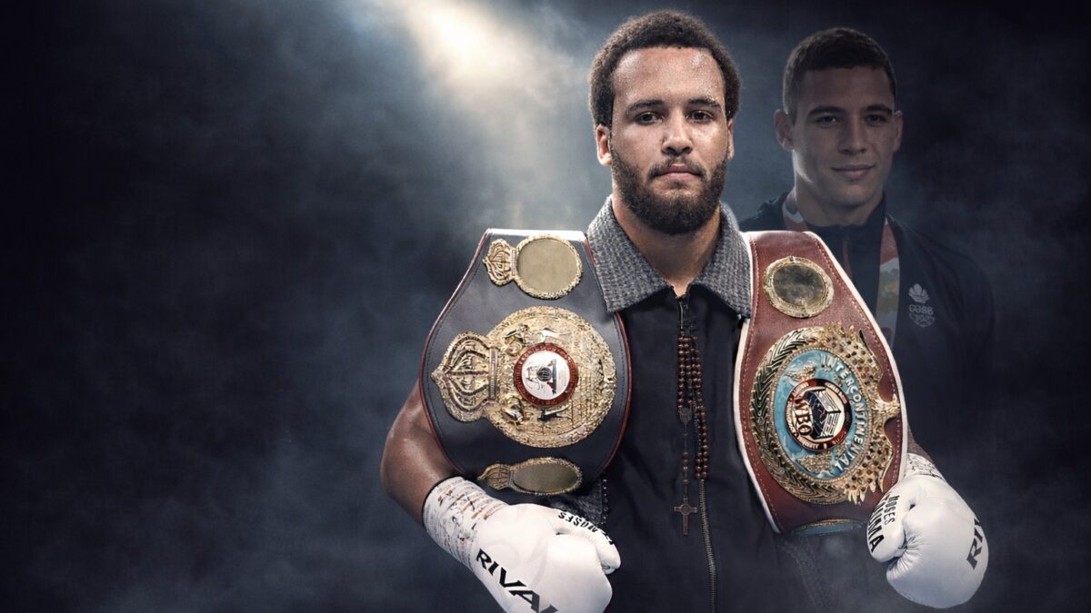 Moses Itauma stands in the ring wearing championship belts, with his brother Karol Itauma visible in the background under subdued lighting.
